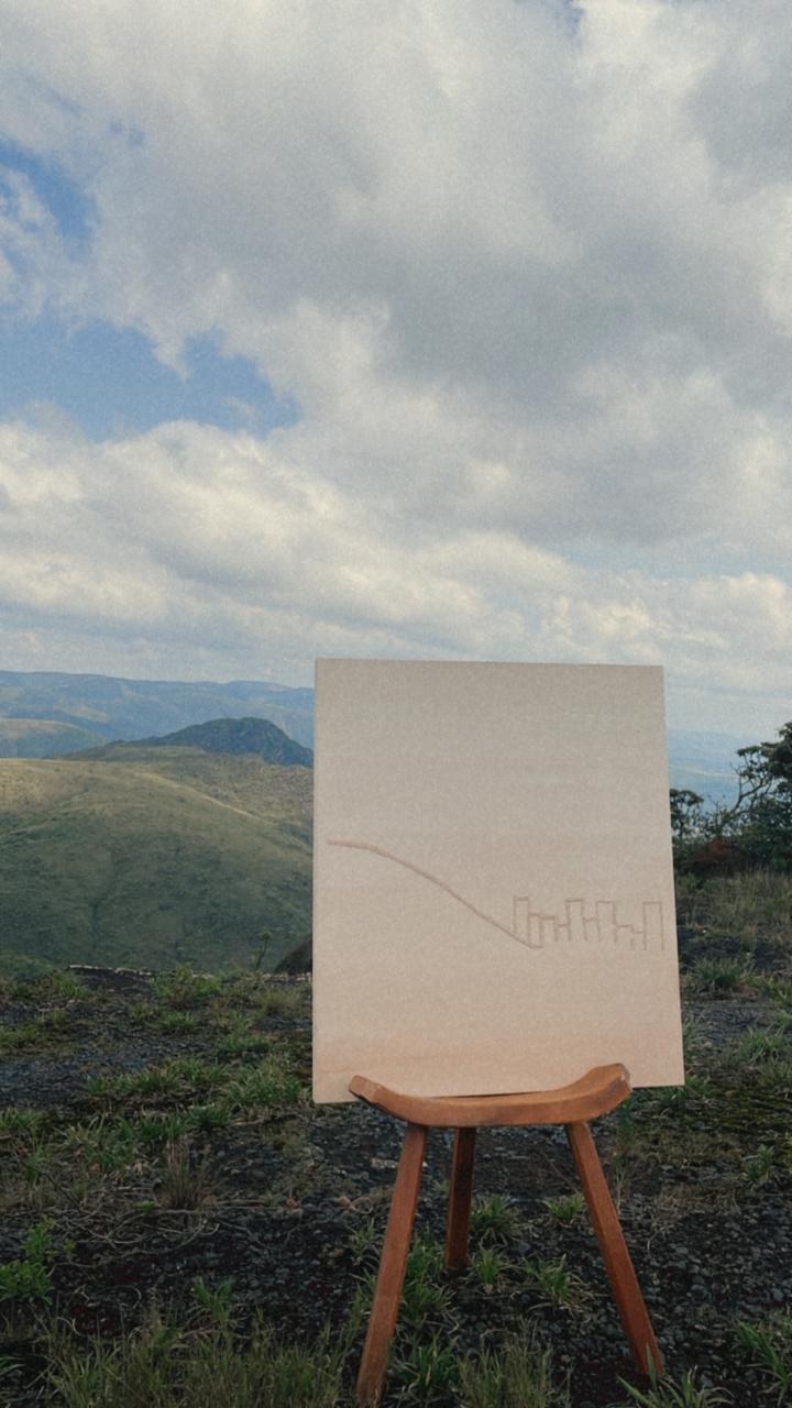 Tela de Vi Cunha apoiada em um banco, diante de uma paisagem montanhosa.
